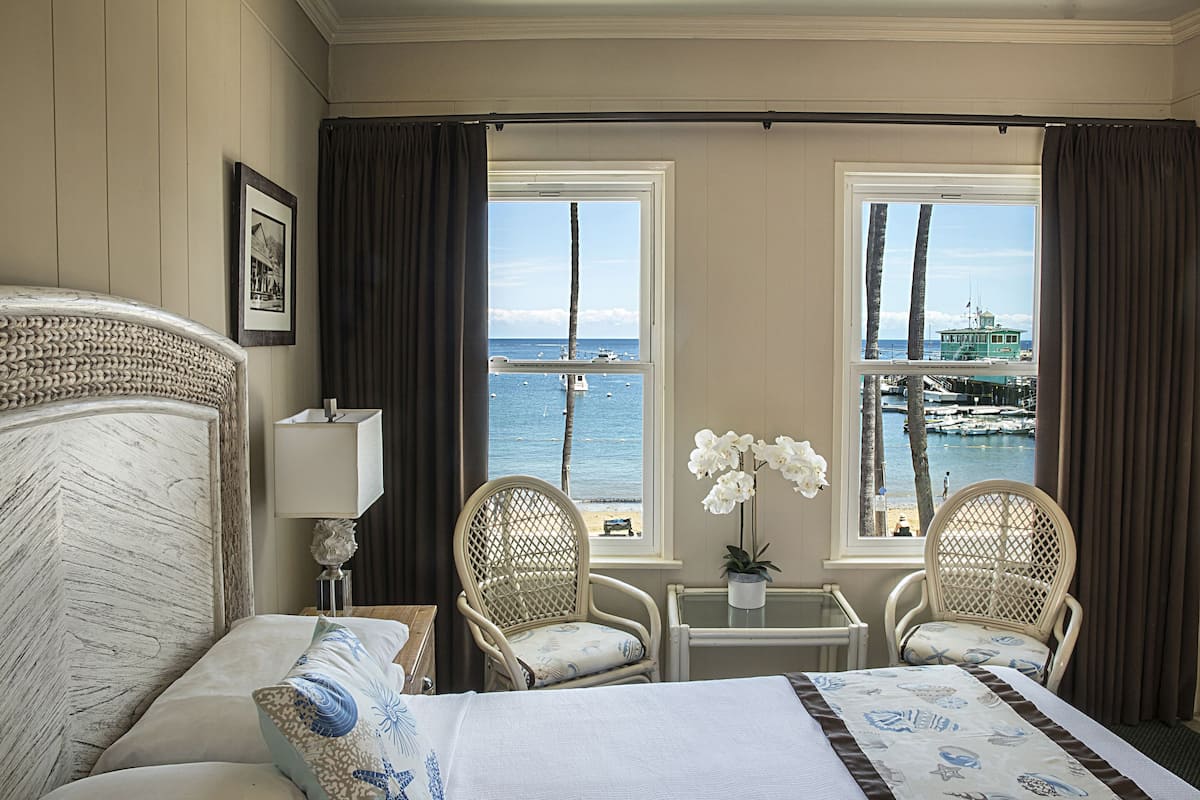 Habitación de hotel con cama, dos sillones de mimbre, una mesita con lámpara y vistas al mar y a los barcos a través de dos ventanas.