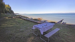A picnic table with a patterned tablecloth and benches, set on a grassy area by a body of water with a clear sky. - Ocean View Tent Site T12 (Port Angeles)