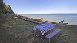 A picnic table with a patterned tablecloth and benches, set on a grassy area by a body of water with a clear sky.
