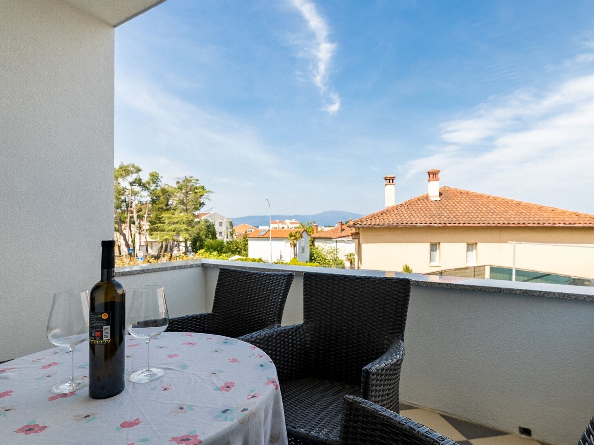 A balcony with a table, two wicker chairs, a bottle of wine, and wine glasses.