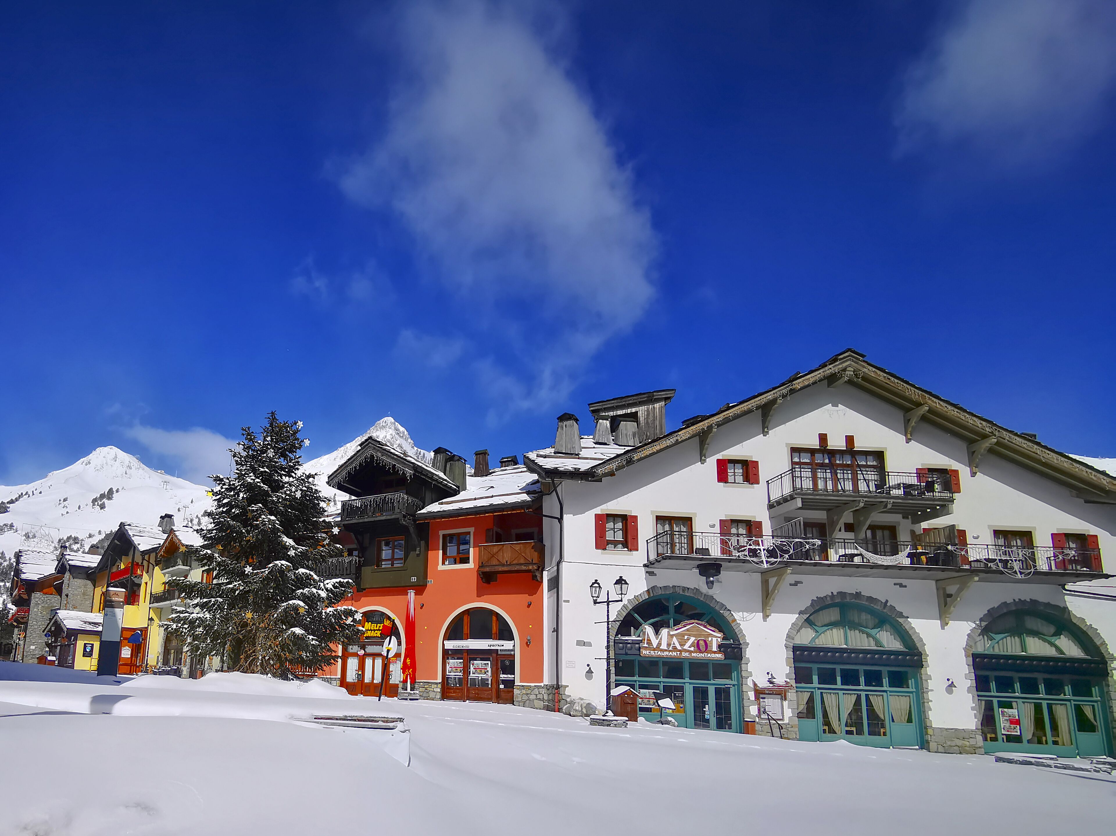 A snow-covered mountain town with colorful buildings, including a restaurant named 'Mazot.'