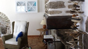 Une pièce avec une cheminée en pierre, un fauteuil en osier, une petite table et une fenêtre ornée d’un motif en forme de croix.
