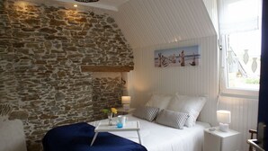 Une chambre avec un mur en pierre, un lit avec une literie blanche, une table de chevet avec une lampe et une fenêtre donnant sur la mer.