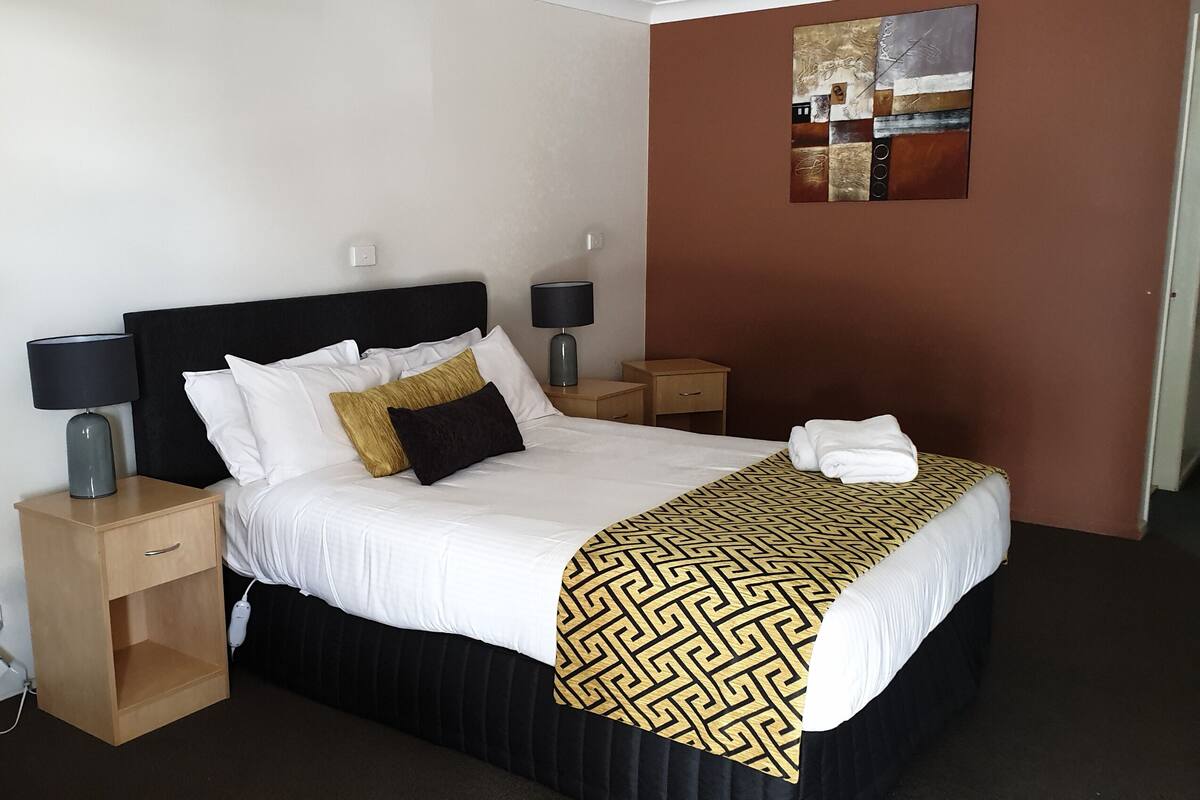 Una cama bien tendida con cabecera negra, ropa de cama blanca y un plaid estampado. Hay dos mesitas de noche con lámparas, un tocador de madera con cajón y una obra de arte en la pared sobre la cama.