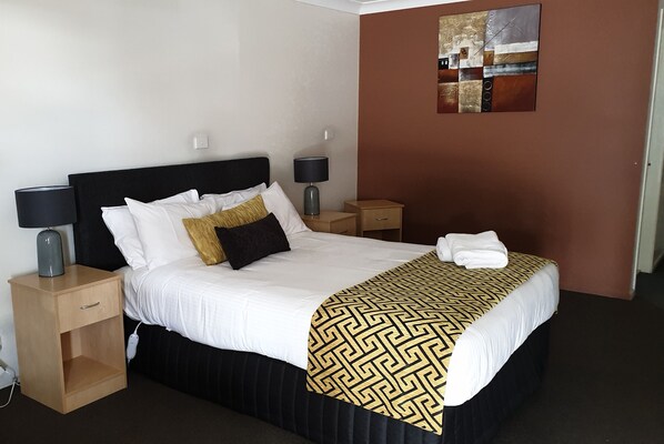 A neatly made bed with a black headboard, white bedding, and a patterned throw. There are two bedside tables with lamps, a wooden nightstand with a drawer, and a wall art piece above the bed.