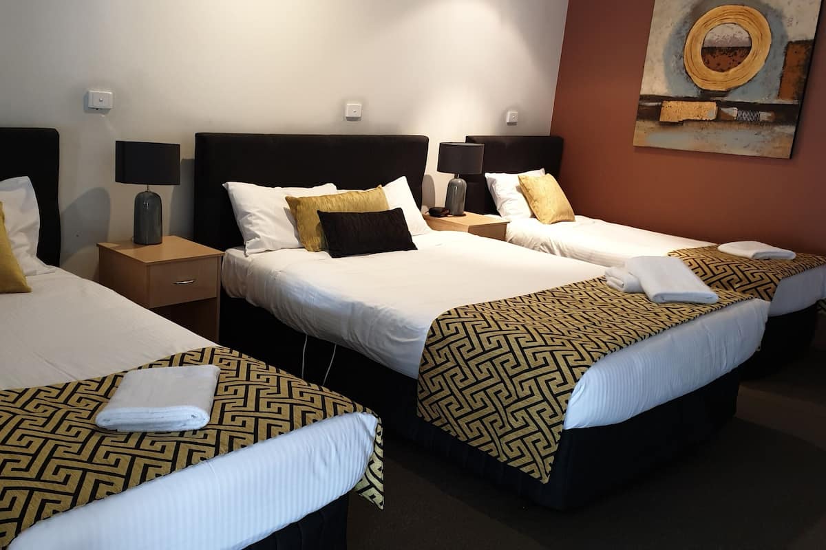 Habitación de hotel con tres camas, cada una con una colcha estampada, una mesita de noche con lámpara y una obra de arte colgada en la pared.