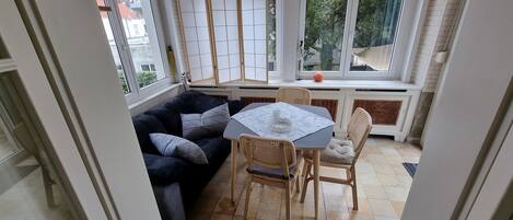 Una habitación acogedora con un sofá azul oscuro, una pequeña mesa redonda, sillas de mimbre y vistas amplias a través de grandes ventanales.