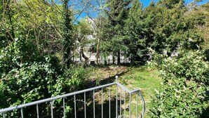 A pathway surrounded by dense greenery and trees, leading to a fenced area with a building in the background.