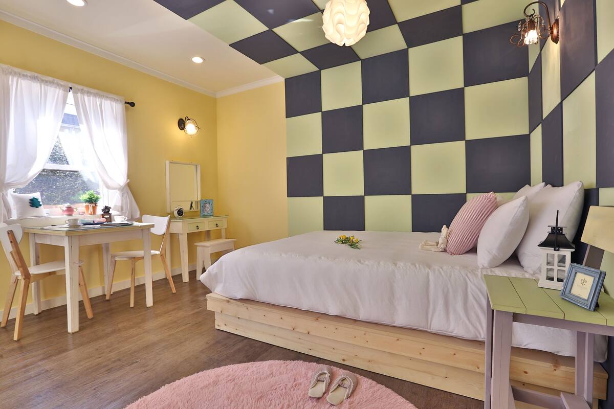 Une chambre avec un mur à carreaux, un lit avec une literie blanche, un bureau avec des chaises et une petite table avec une lampe.