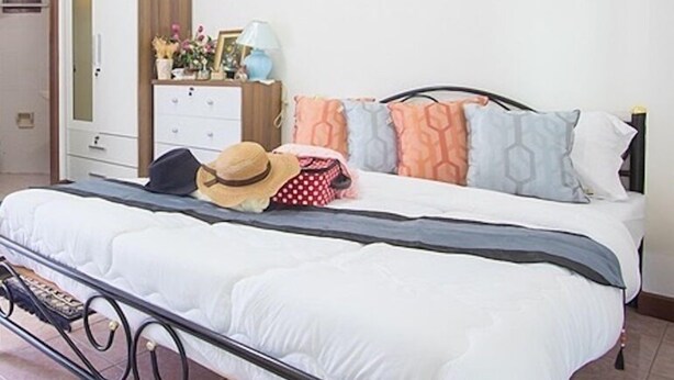 A neatly made bed with a white comforter and a metal frame, a wooden nightstand with a lamp and decorative items, and a wardrobe with a mirror. - P Apartment Nonthaburi (Nonthaburi)