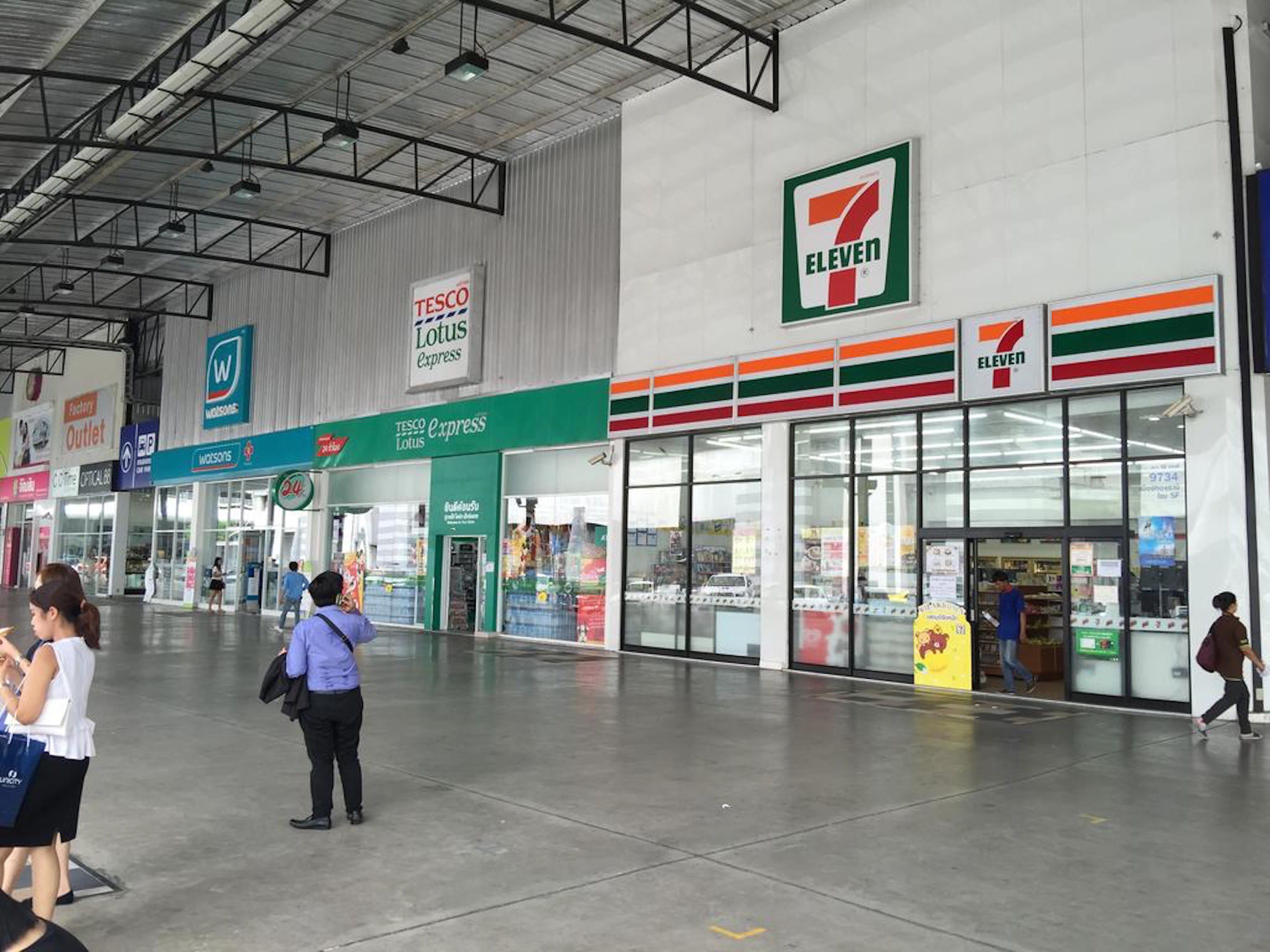 A 7-Eleven store with a green and white sign, a Tesco Express, and a Lotus Express inside a large indoor shopping area.