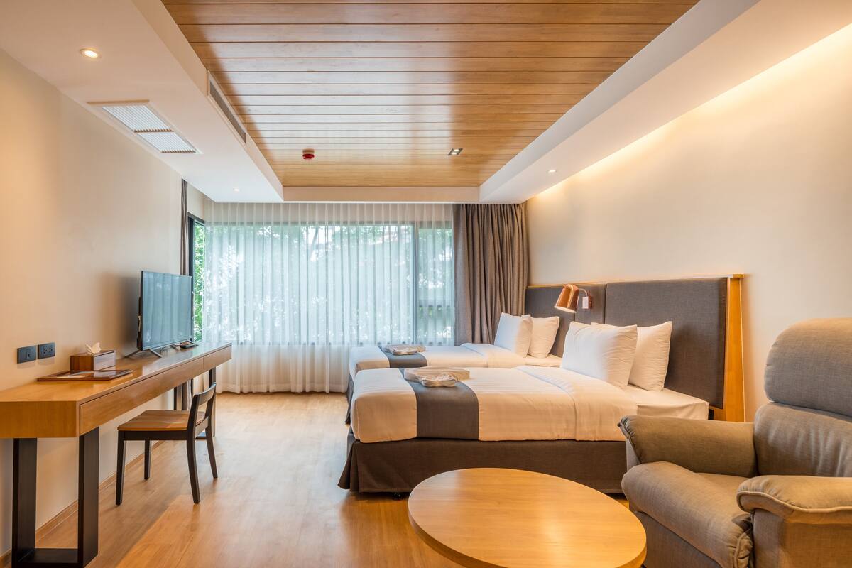 A hotel room with a wooden ceiling, a desk with a computer, two beds, a round coffee table, and a sofa.