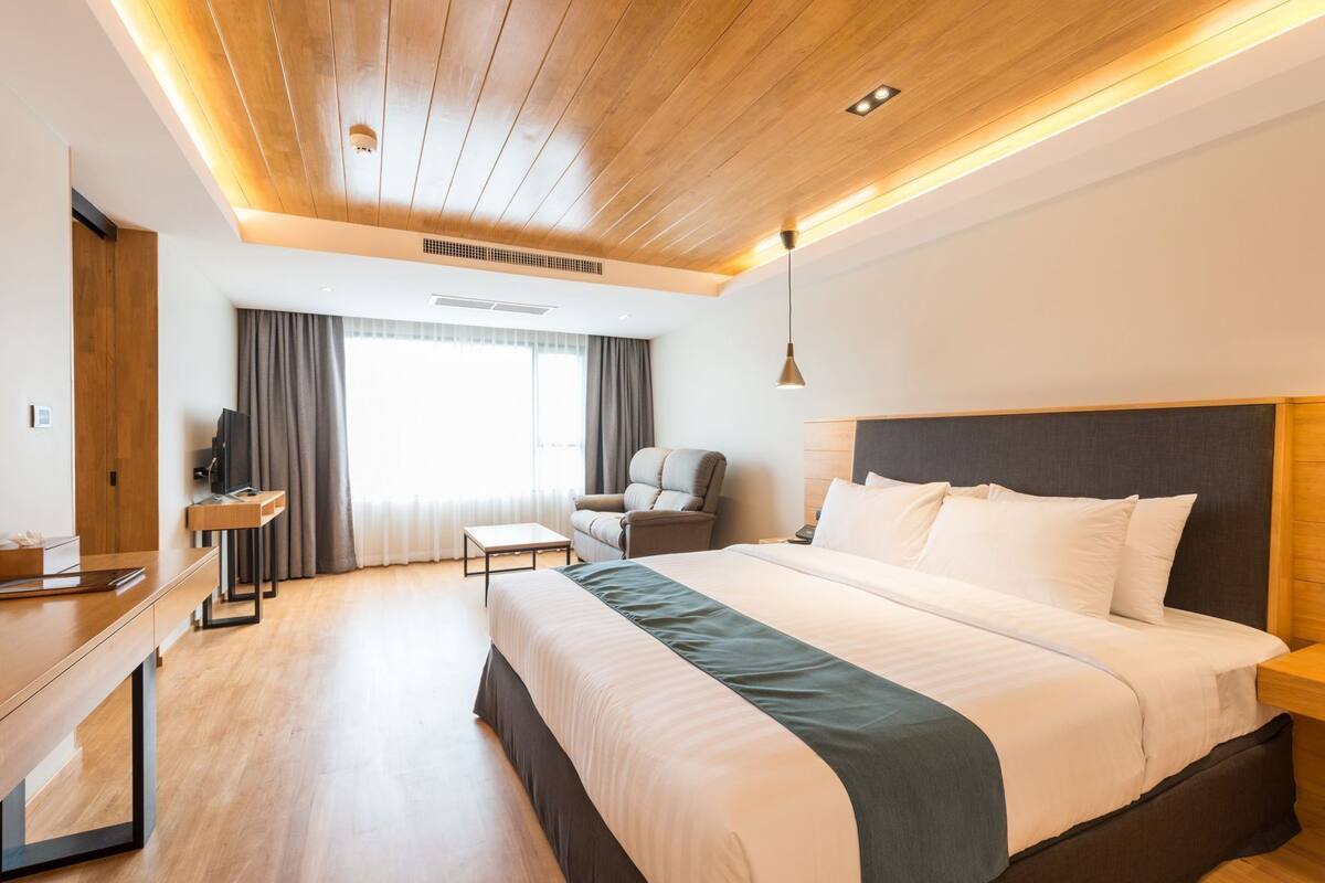 A modern hotel room with a wooden ceiling, a large bed, a desk, a chair, and a TV.