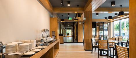 A modern restaurant with wooden beams, a long counter with stacked plates, and a view through large windows.