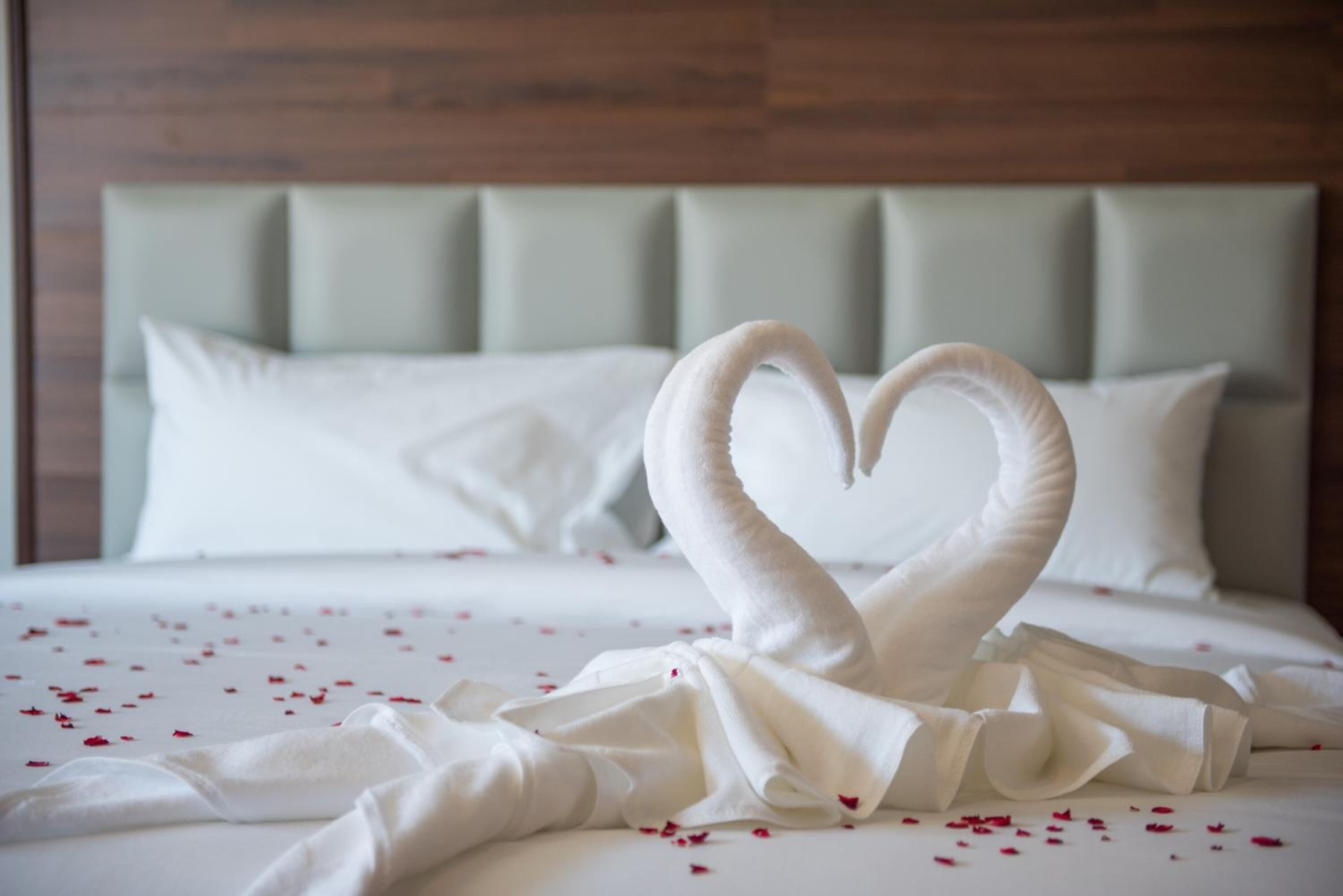 A bed with a heart-shaped towel arrangement and scattered rose petals.