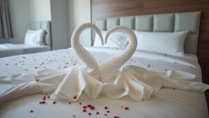 Una habitación de hotel con una cama decorada con una toalla en forma de corazón y pétalos de rosa esparcidos.