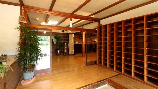 Ett traditionellt japanskt rum med trägolv, en stor krukväxt, en bokhylla i trä och en skjutdörr som leder till ett annat rum.
