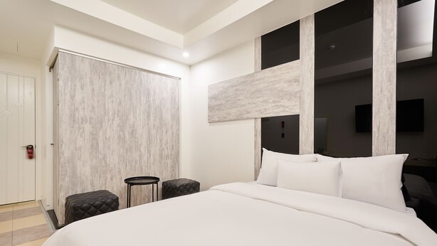 Una habitación de hotel moderna con una cama grande, un armario, un taburete pequeño y un televisor montado.