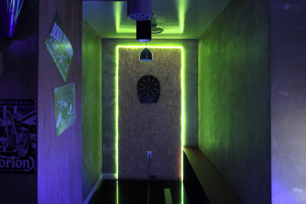 A narrow hallway with a dartboard mounted on the wall, green neon lighting, and a ceiling light fixture.