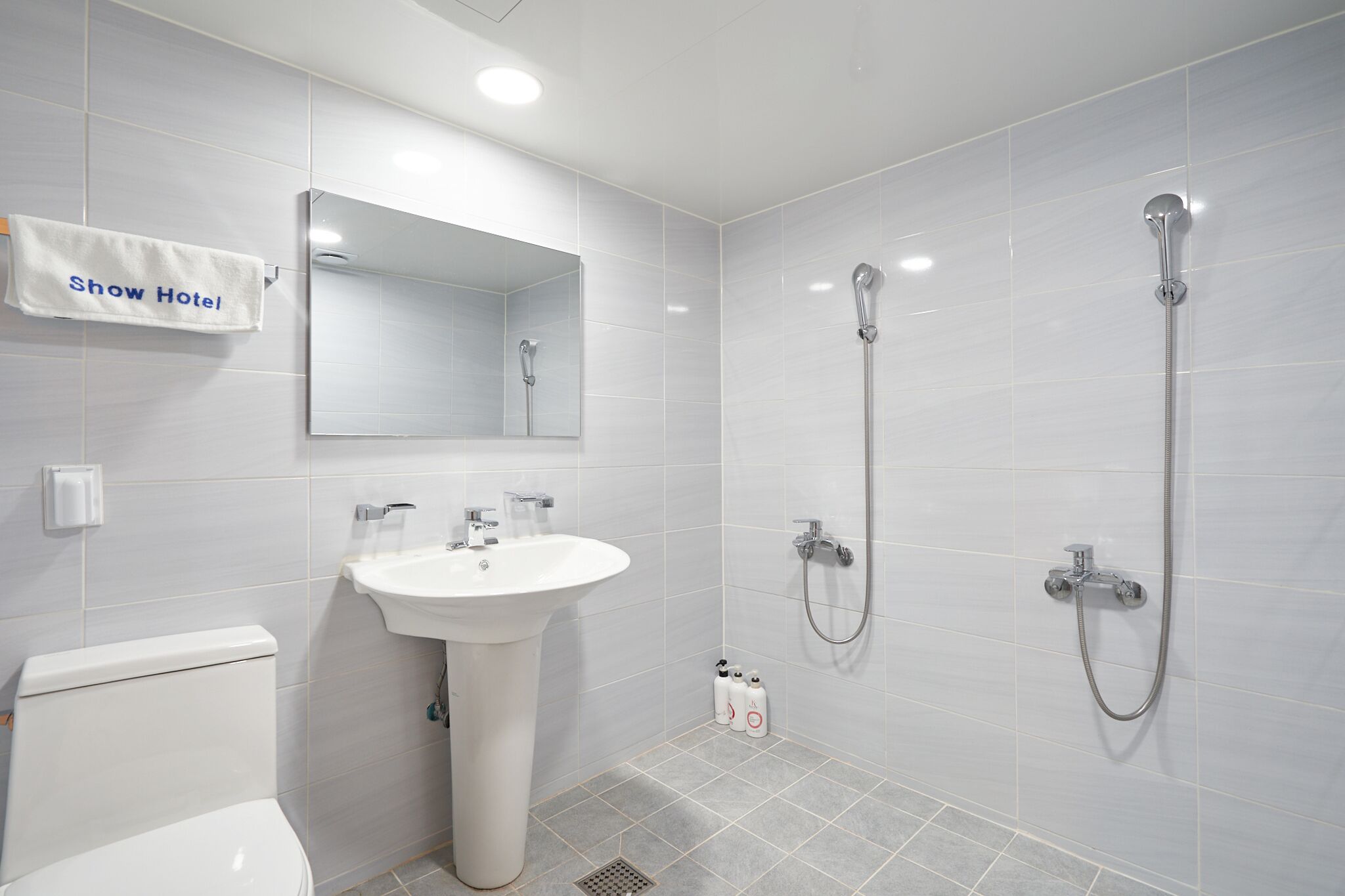 Une salle de bain moderne avec un lavabo blanc, deux douches, des toilettes et un porte-serviettes sur lequel est accroché un panneau indiquant « Show Hotel ».