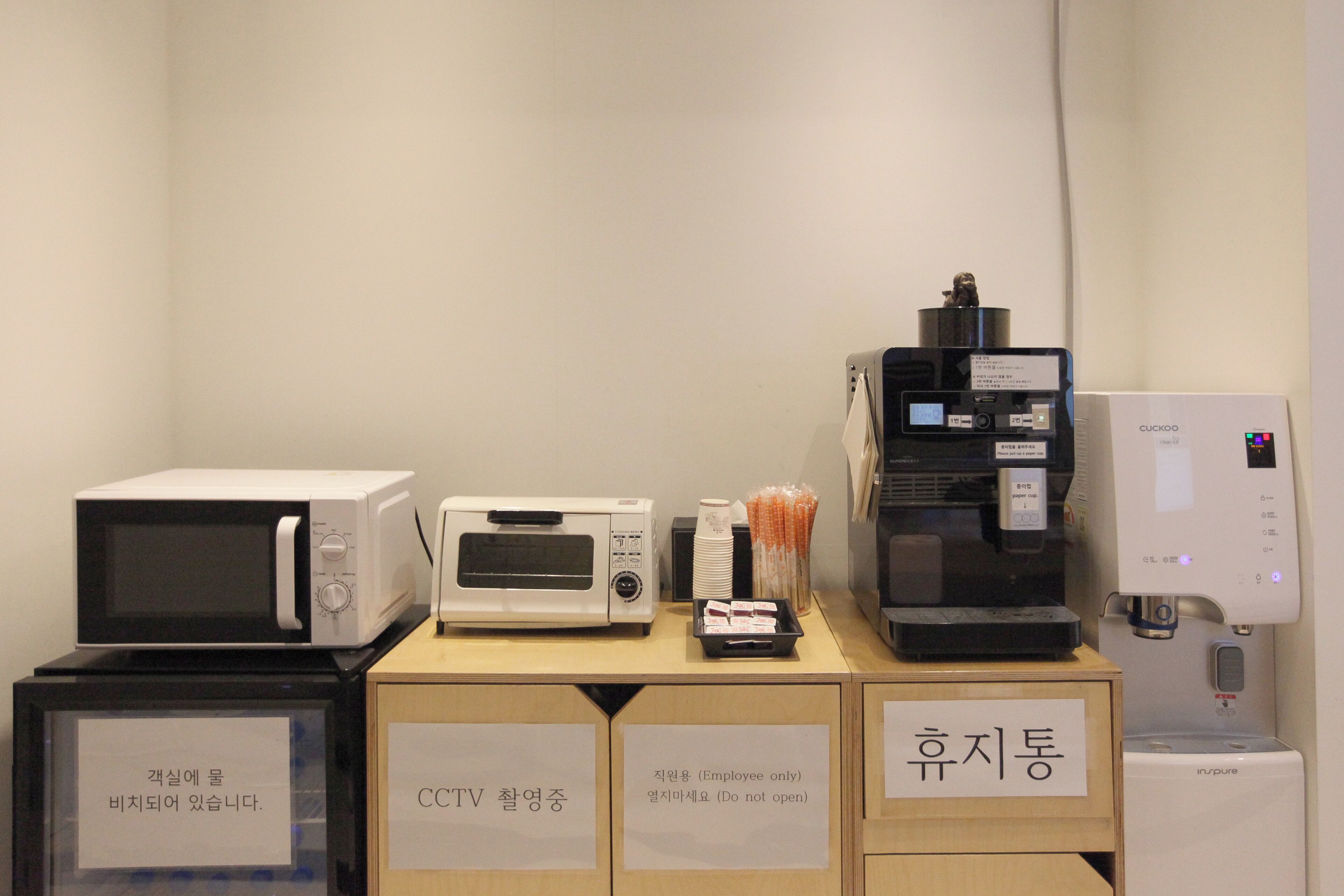 Eine Küchenzeile mit Mikrowelle, Toaster und Kaffeemaschine. Es sind Schilder für Mitarbeiterbereiche angebracht und es gibt einen Hinweis zur Kaffeemaschine.