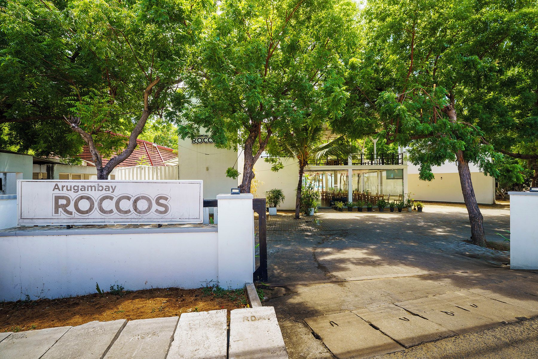 A sign for Arugambay Roccos, a paved pathway, and a building with a balcony.