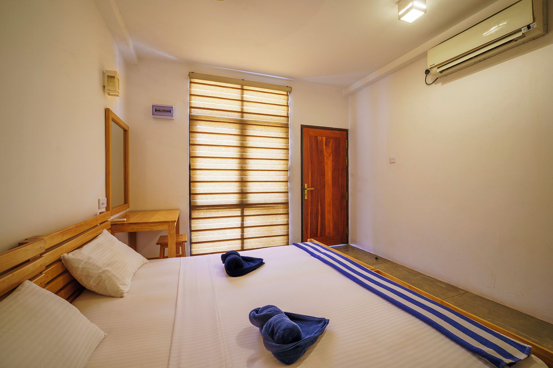 A hotel room with a bed, a wooden headboard, a bedside table, a mirror, a window with blinds, a door, and an air conditioning unit.