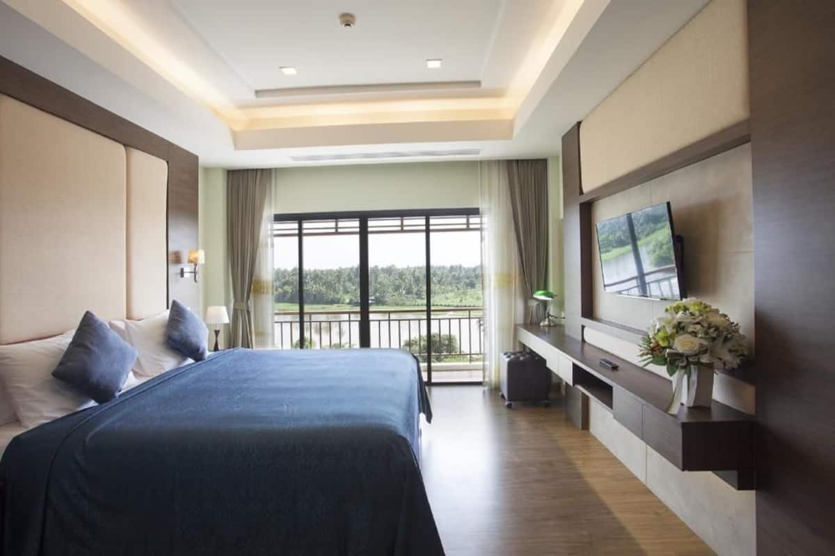 A hotel room with a large bed, a TV mounted on the wall, a balcony with a view, and a desk with a vase of flowers.