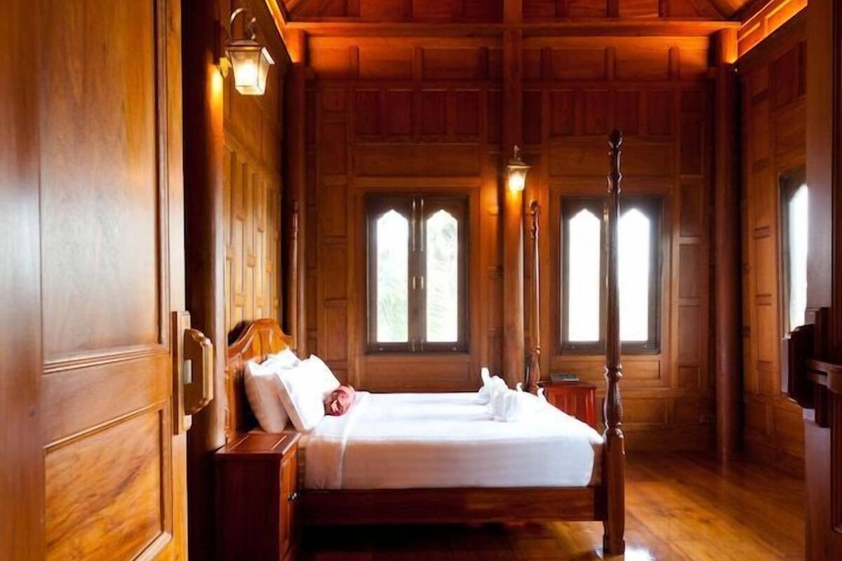 A wooden four-poster bed in a room with wooden walls and floors, two windows, and wall-mounted lights.