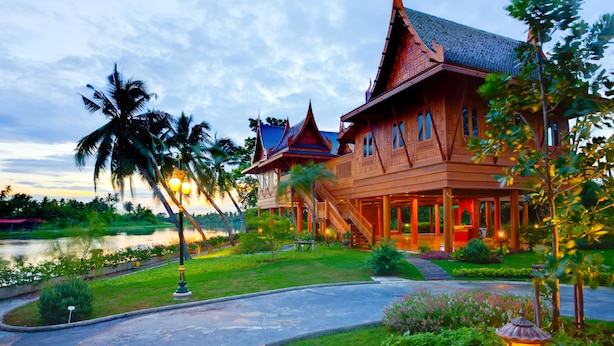 A traditional wooden building with multiple levels and a steeply pitched roof, surrounded by lush greenery and a body of water. - RK Riverside Resort & Spa (Sam Phran)