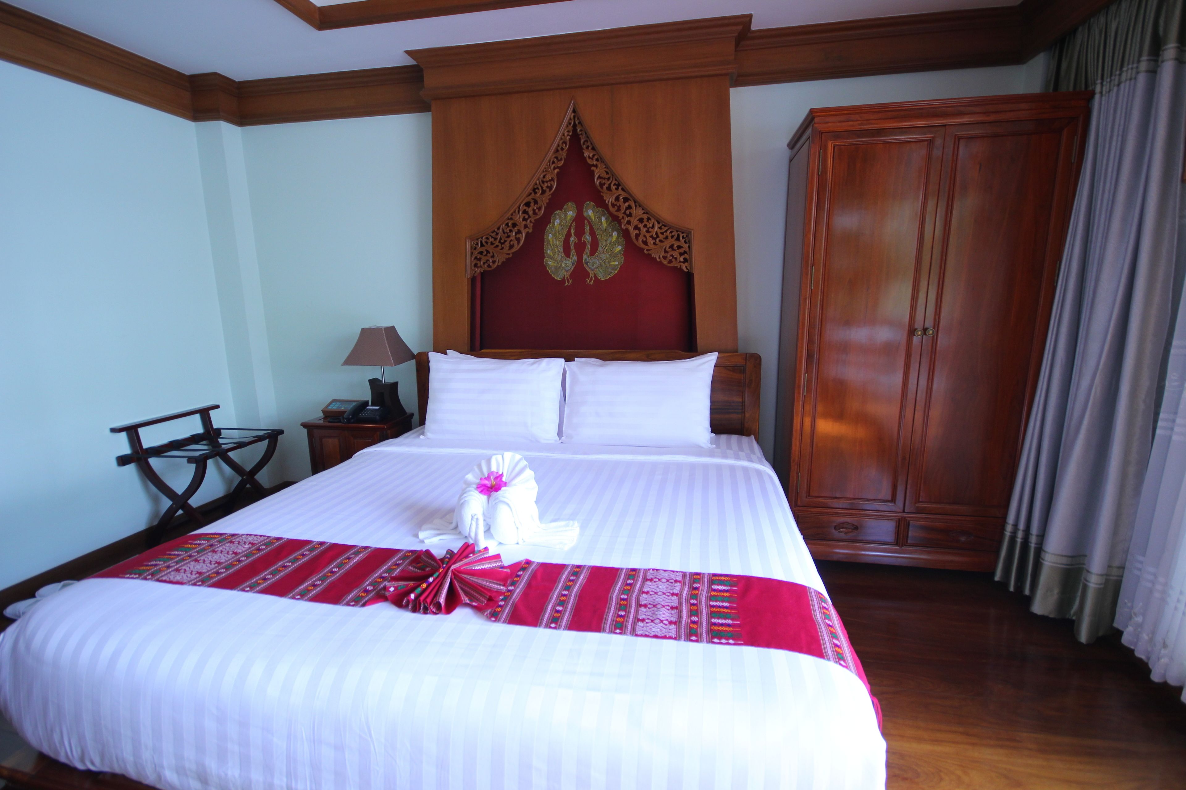 A neatly made bed with white linens, a red and white patterned bedspread, and a white towel with a pink bow. There is a wooden headboard with a decorative design, a wooden wardrobe, and a small table with a lamp beside the bed.