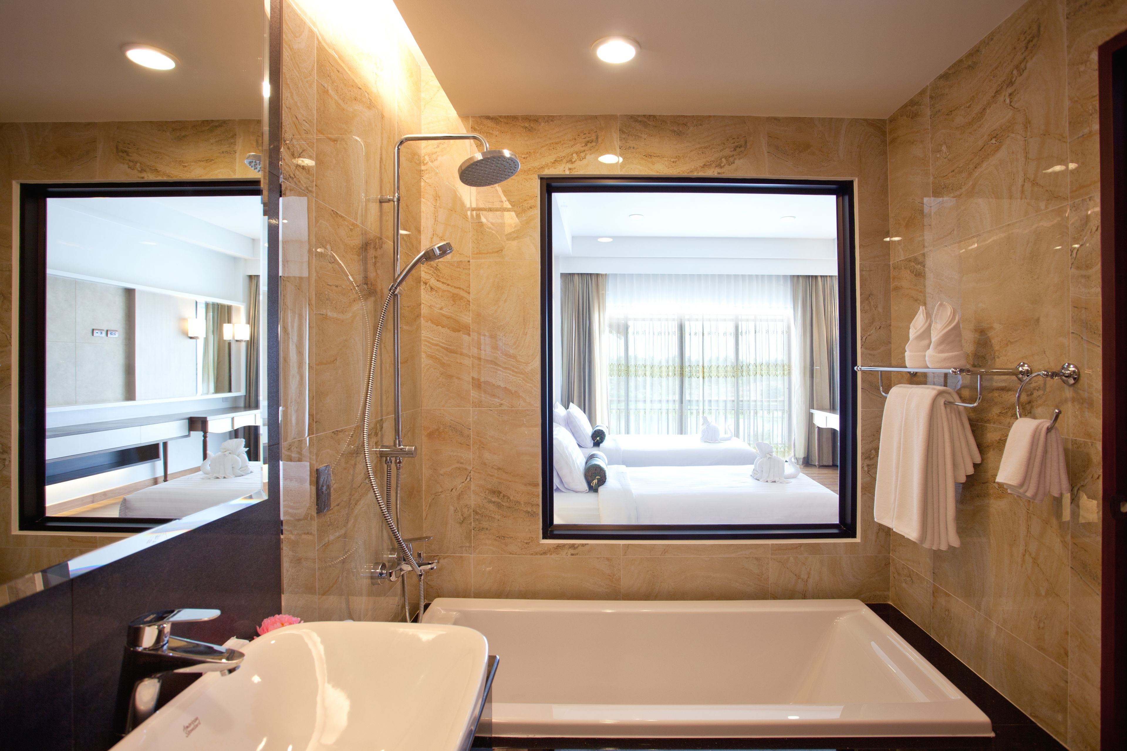 A modern bathroom with a large bathtub, a shower area, and a mirror reflecting a bedroom with a bed and window.