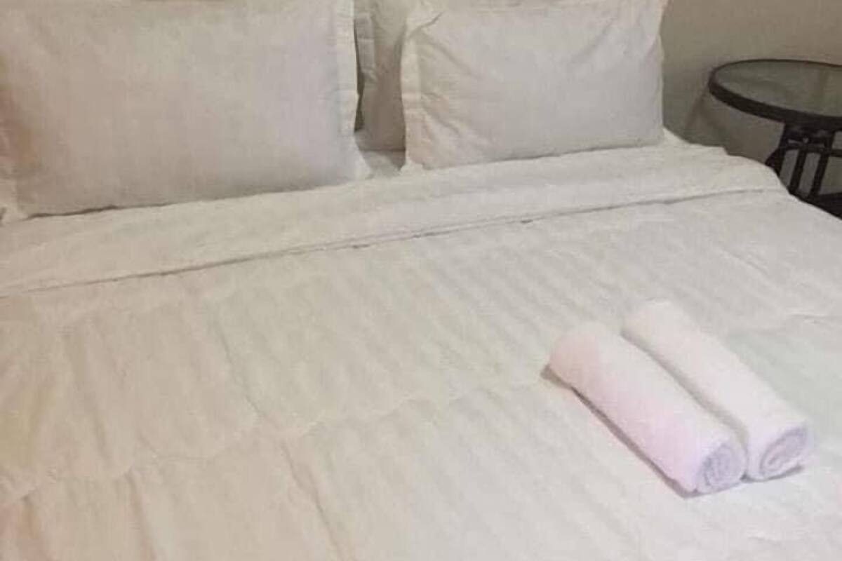 A neatly made bed with white bedding and two pillows, a round table beside it, and a plain wall in the background.