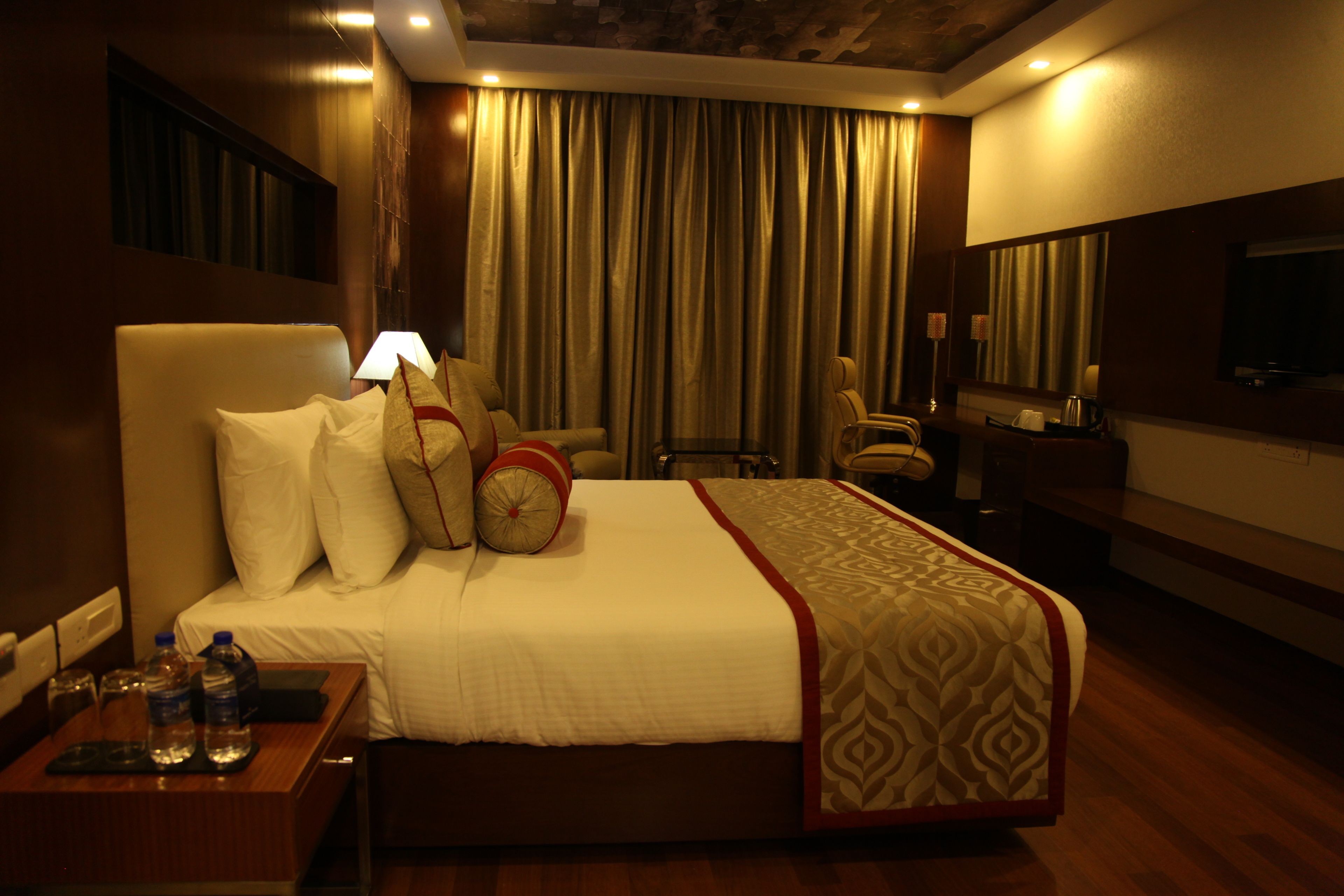A hotel room with a large bed, a desk with a lamp, a chair, a TV, and a window with curtains.