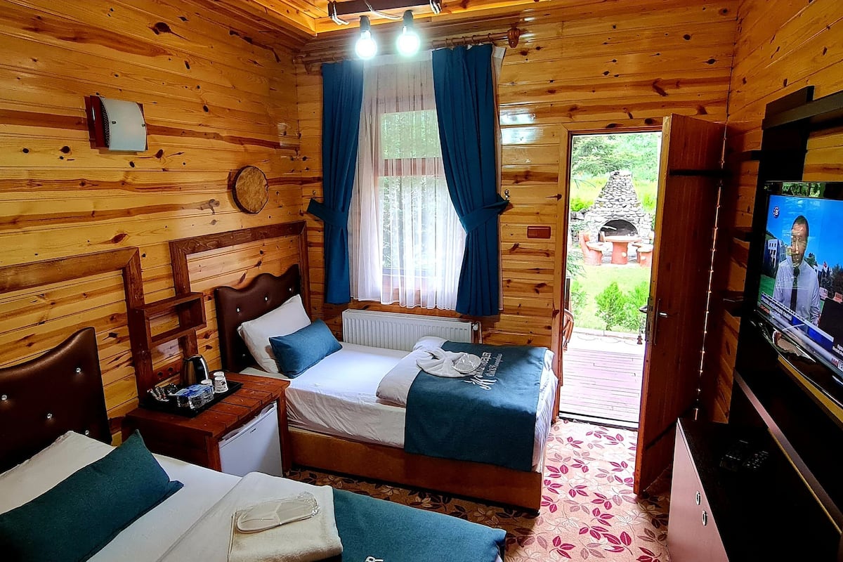 A wooden cabin room with two beds, a small table, a TV, and a window with curtains.