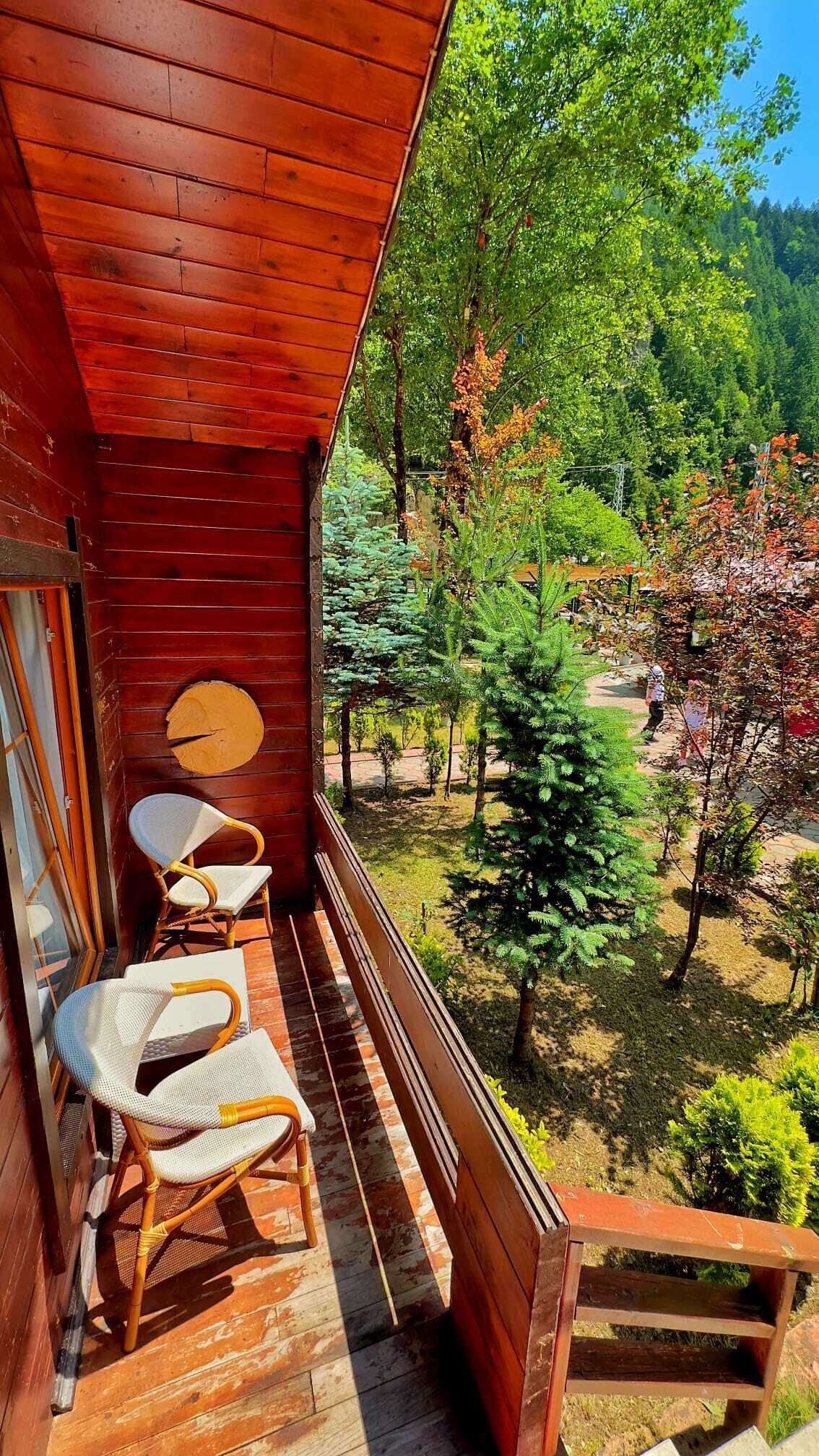 Un balcón de madera con dos sillas de mimbre y una mesa, con vistas a un jardín con árboles y cielo despejado.