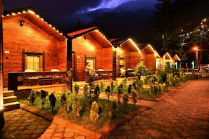 Une rangée de cabanes en bois, éclairées de l’extérieur, un chemin pavé et de petits arbres au premier plan.