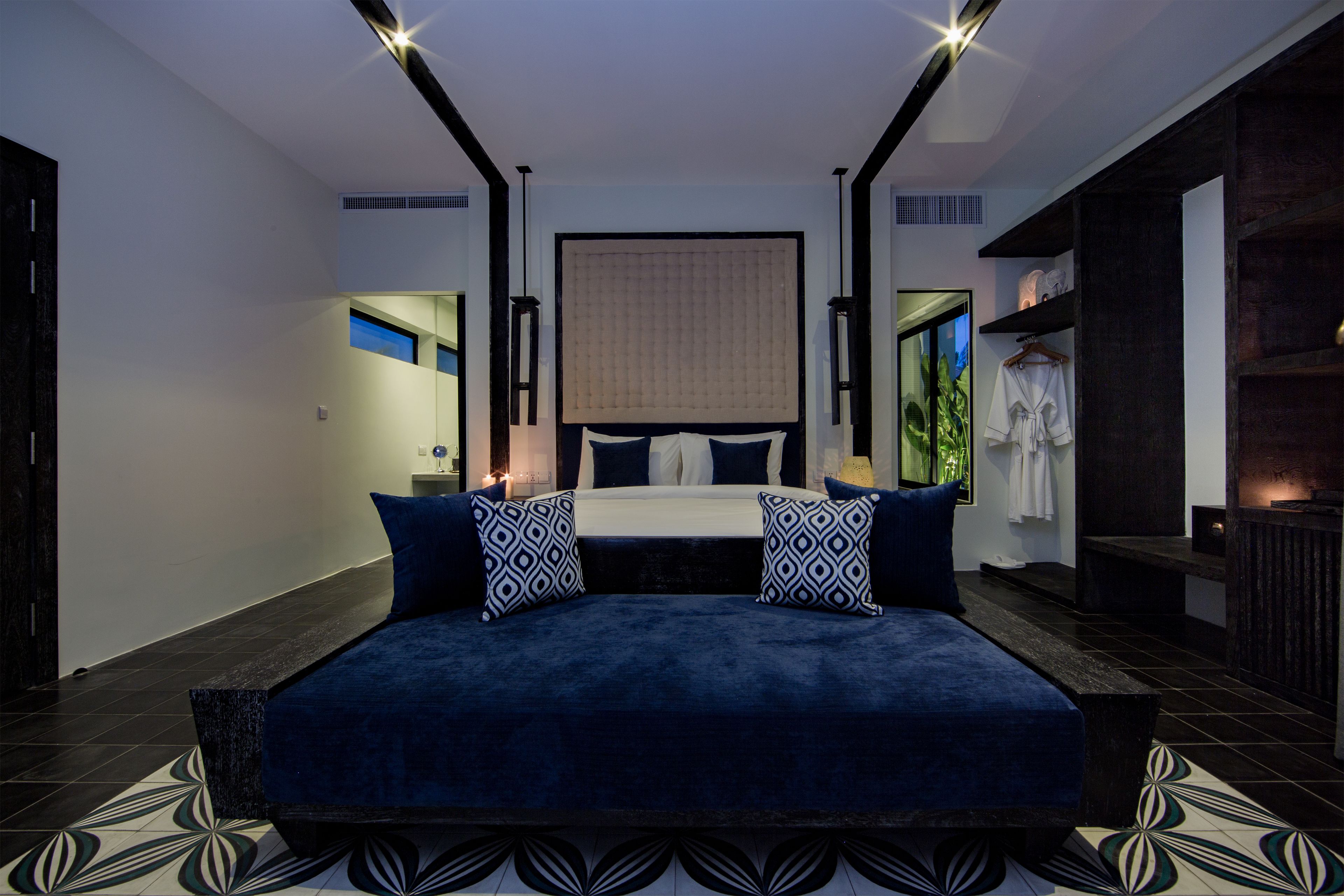 A modern bedroom with a large bed, dark blue bedding, and patterned pillows. There is a nightstand with a lamp, a wardrobe, and a mirror.