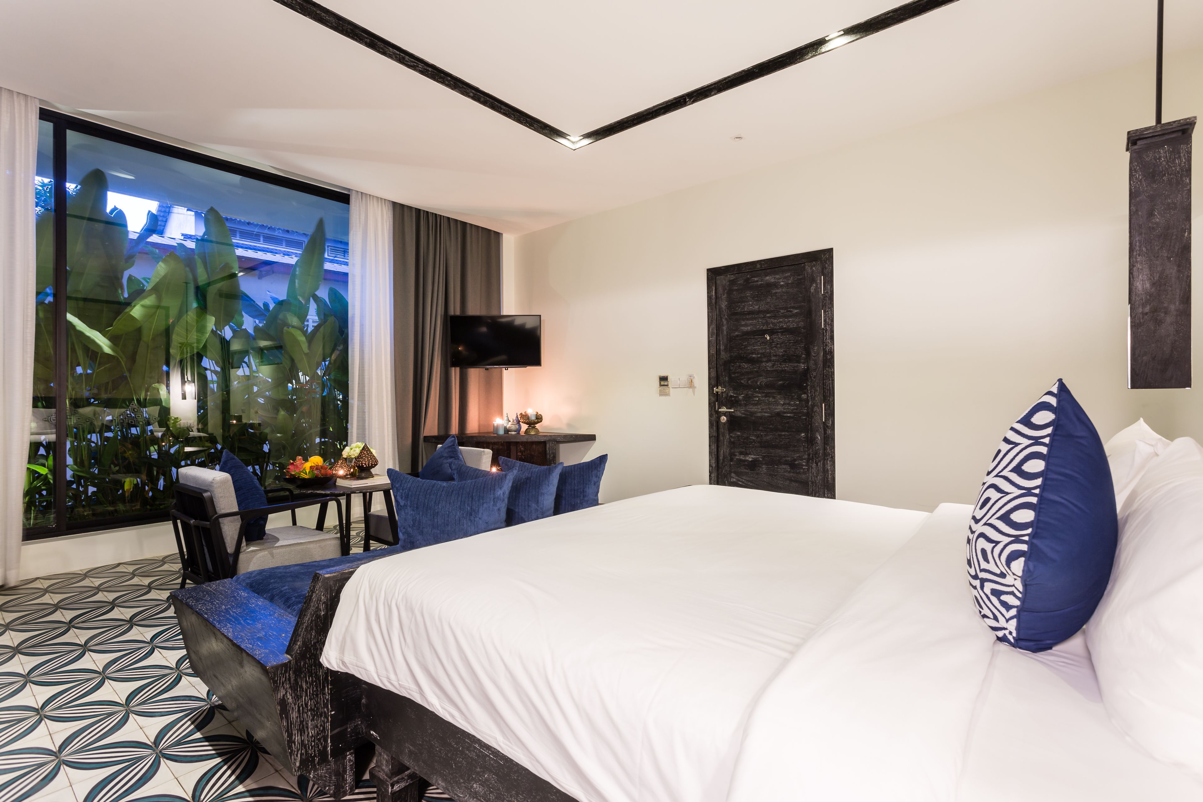 A modern hotel room with a large bed, a flat-screen TV, a dining area with a table and chairs, and a view of greenery through a large window.