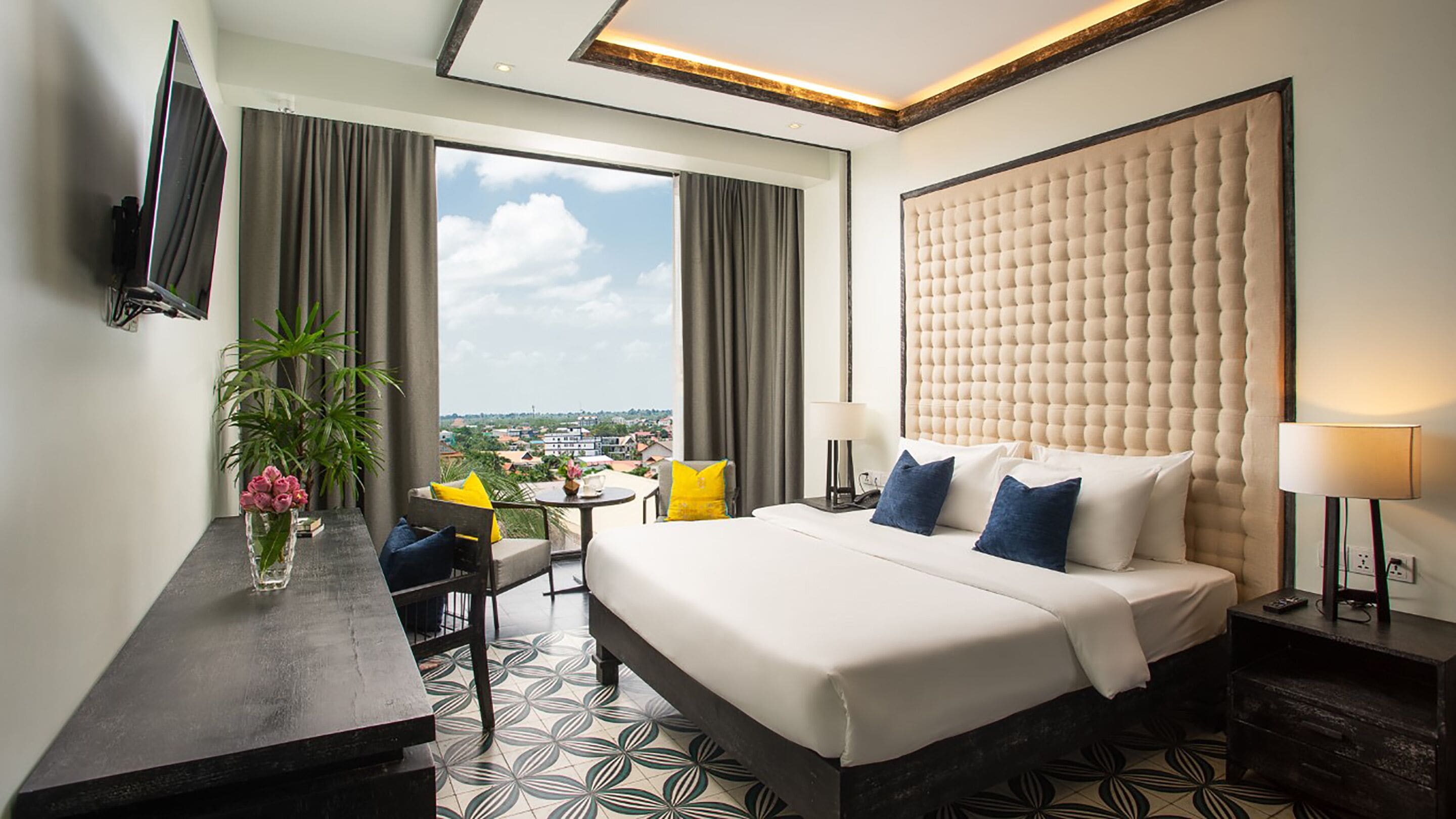 A hotel room with a large bed, a desk, a chair, a TV, and a view of the city.