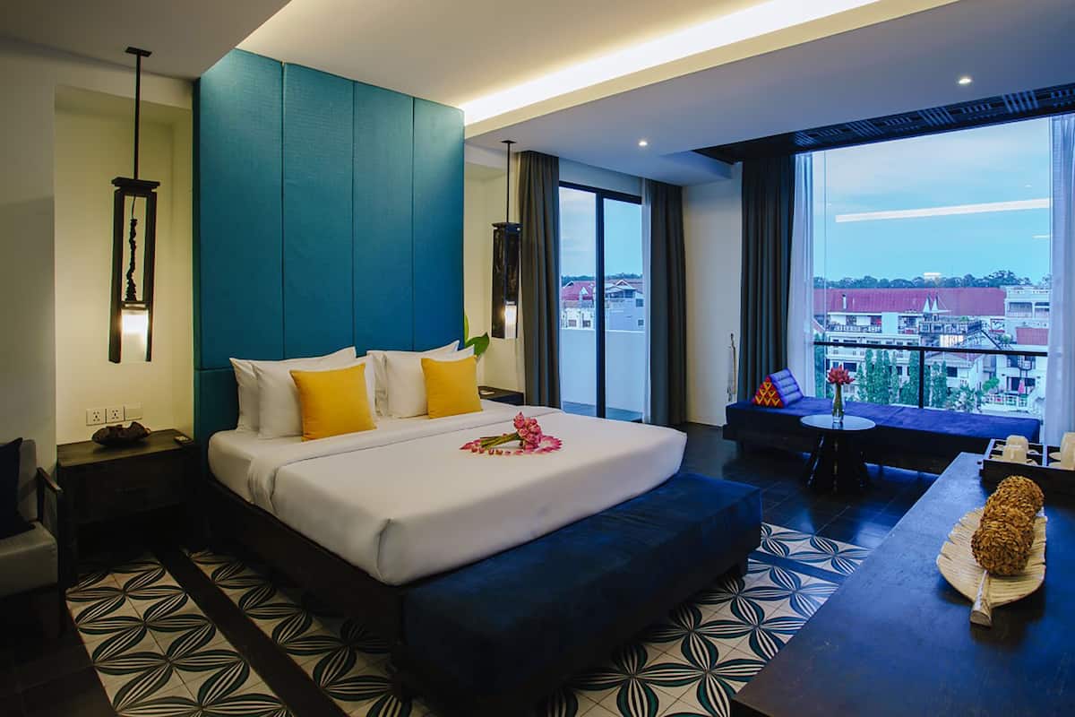 A modern hotel room with a large bed, a sofa, a dining table, and a balcony with a city view.