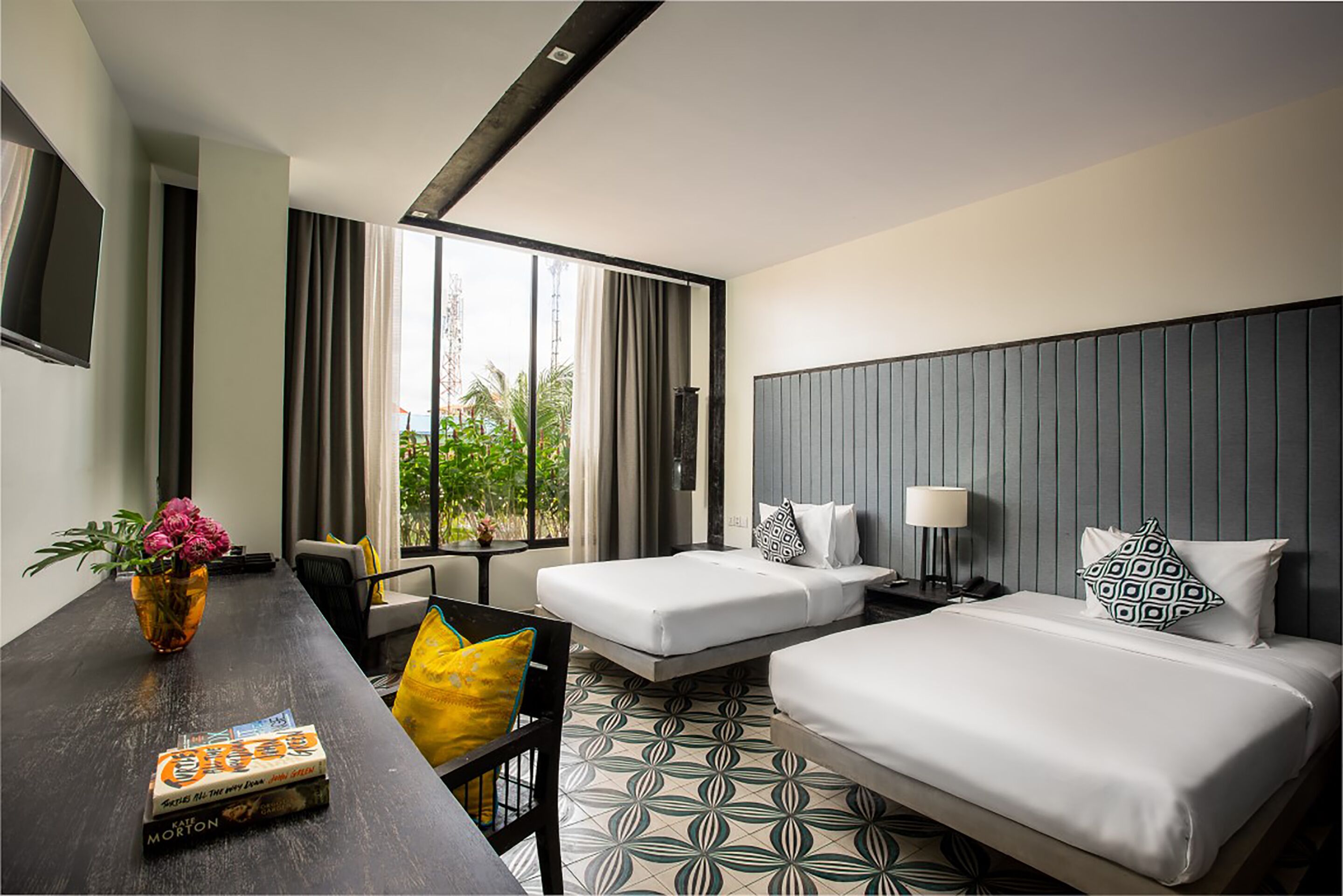 Une chambre d’hôtel avec deux lits, un bureau avec une chaise, un téléviseur fixé au mur et une fenêtre avec des rideaux.