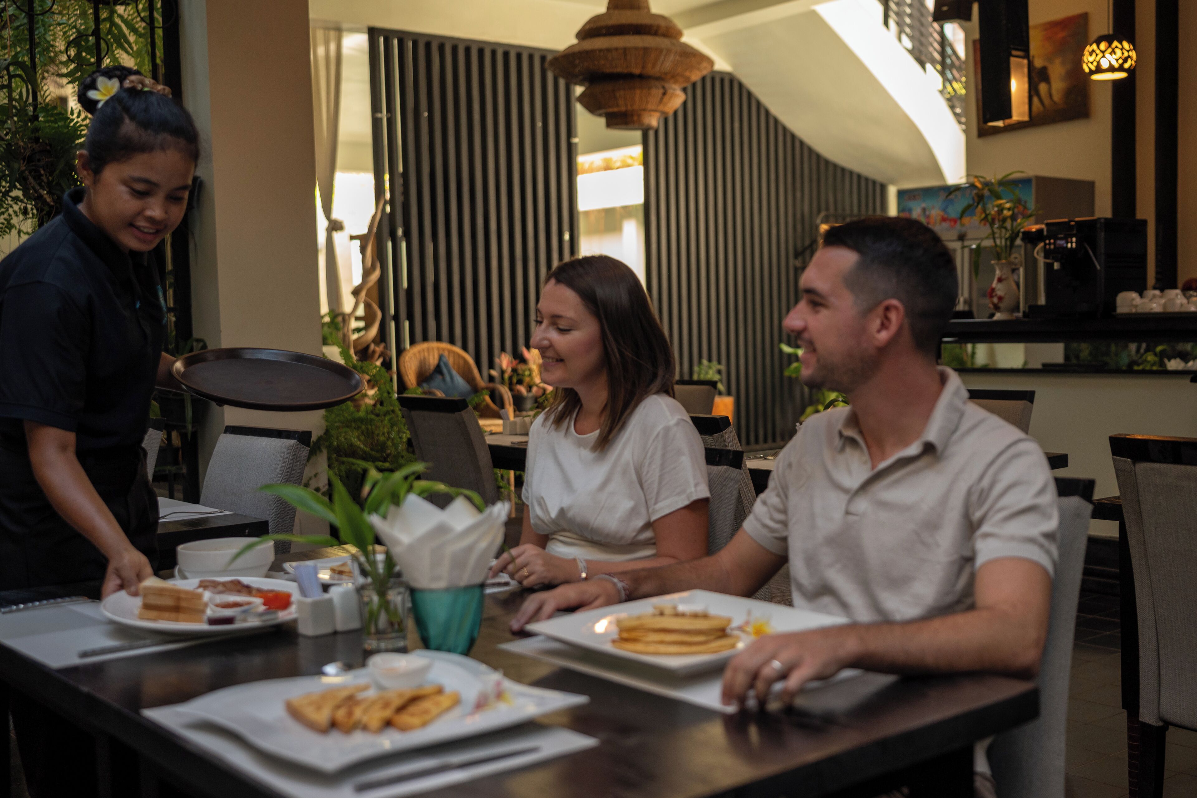 Une salle à manger avec un serveur, un homme et une femme assis à une table où se trouvent des assiettes de nourriture, un vase rempli de fleurs et un luminaire suspendu.