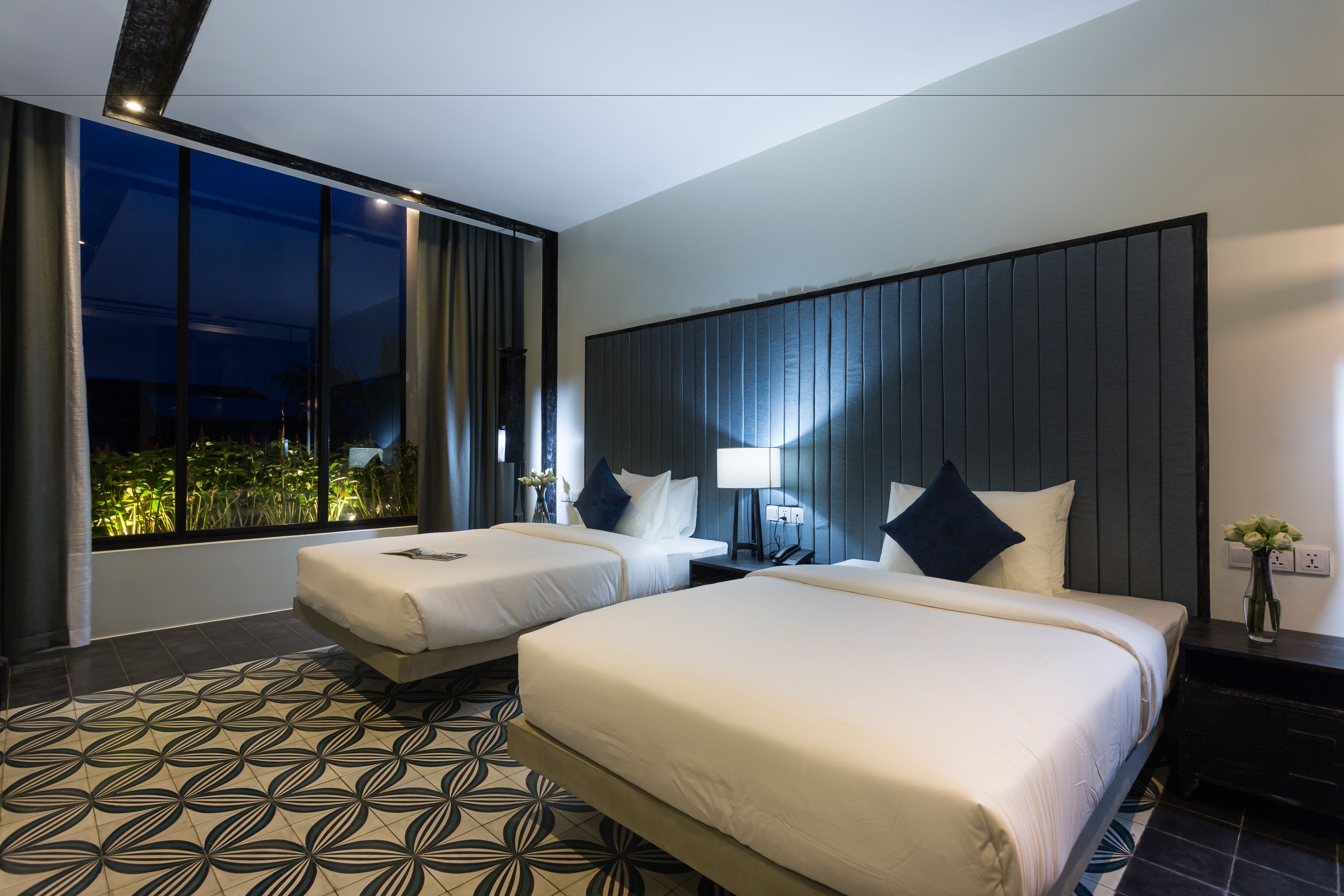 A hotel room with two beds, a large window with a view of greenery, a patterned floor, and a modern design.