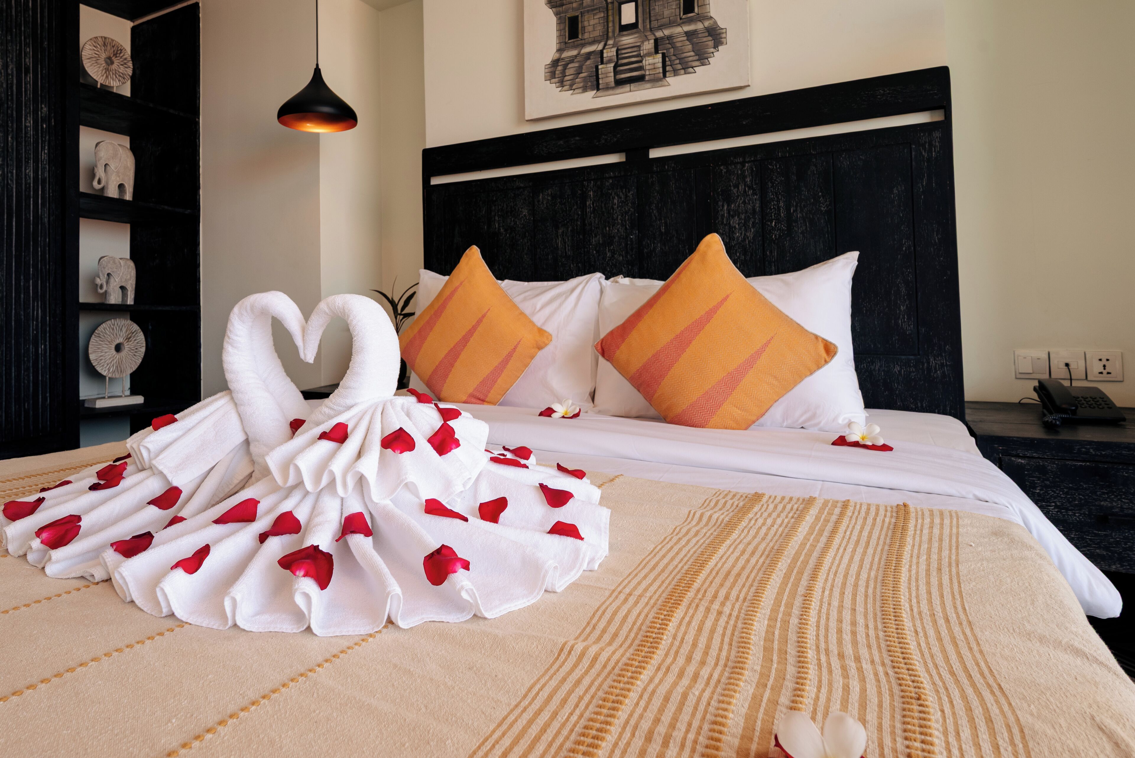 A neatly made bed with a heart-shaped towel arrangement, a black headboard, and a wall-mounted picture.