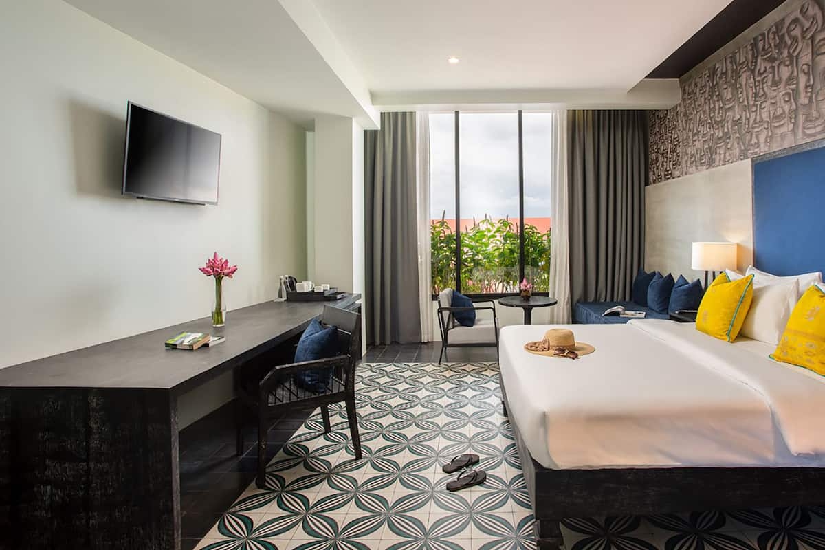 A hotel room with a bed, a desk, a chair, a TV, and a view of greenery outside.
