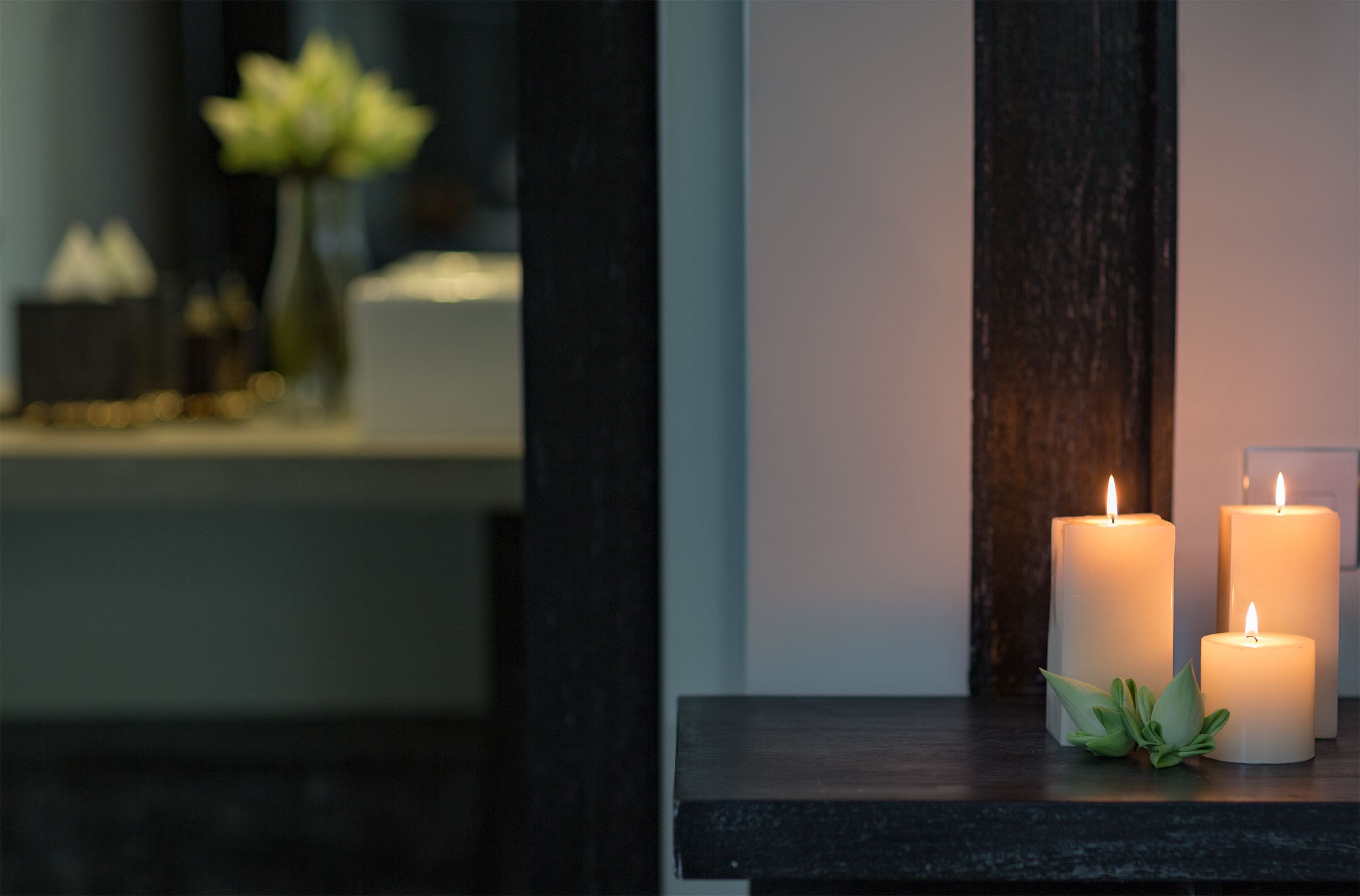 Three lit candles on a dark wooden surface with a vase of white flowers in the background.