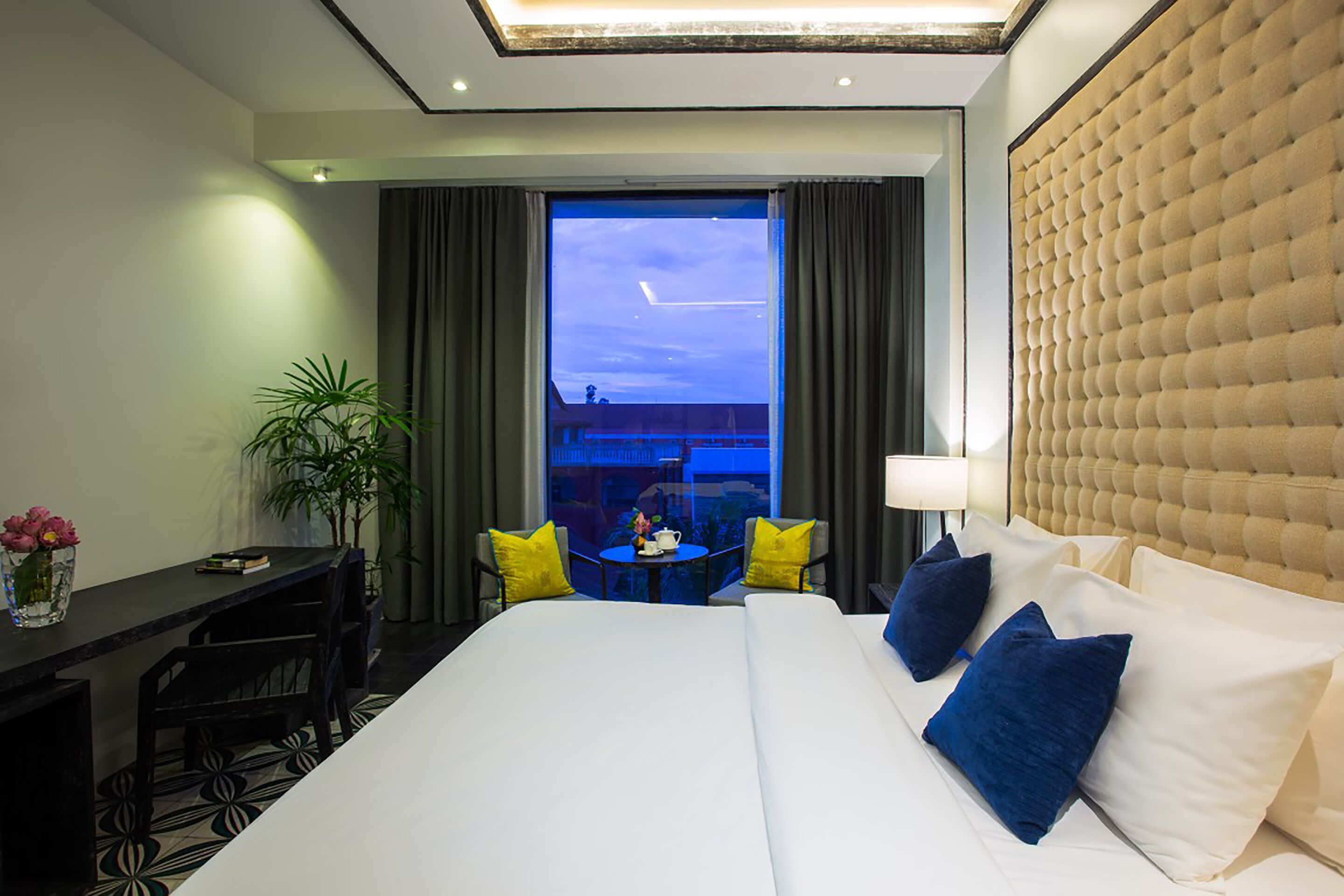 A hotel room with a large bed, a desk, two chairs, a small table, and a potted plant.