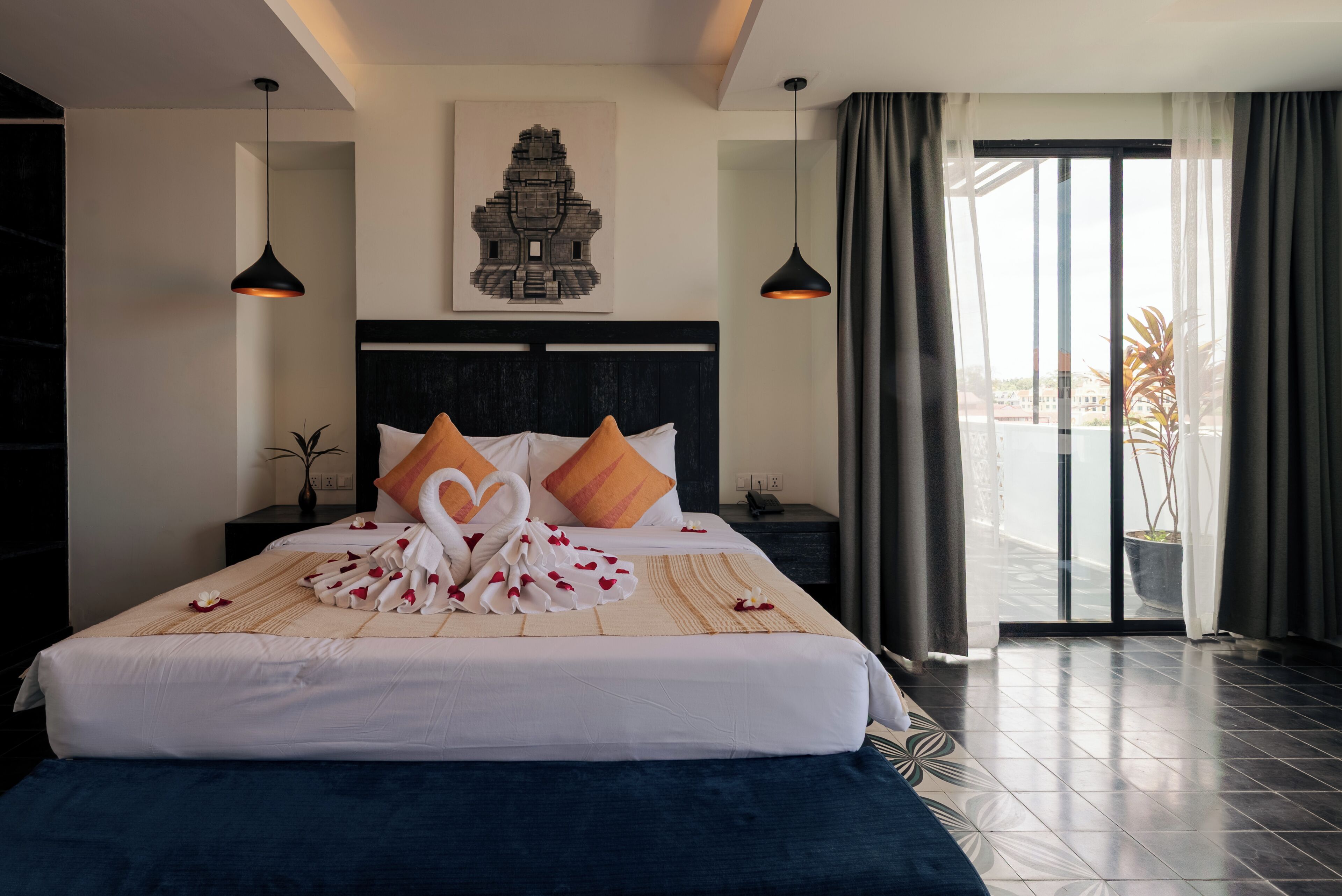 A hotel room with a bed, a headboard, a wall-mounted artwork, a nightstand, a potted plant, and a view through a glass door.