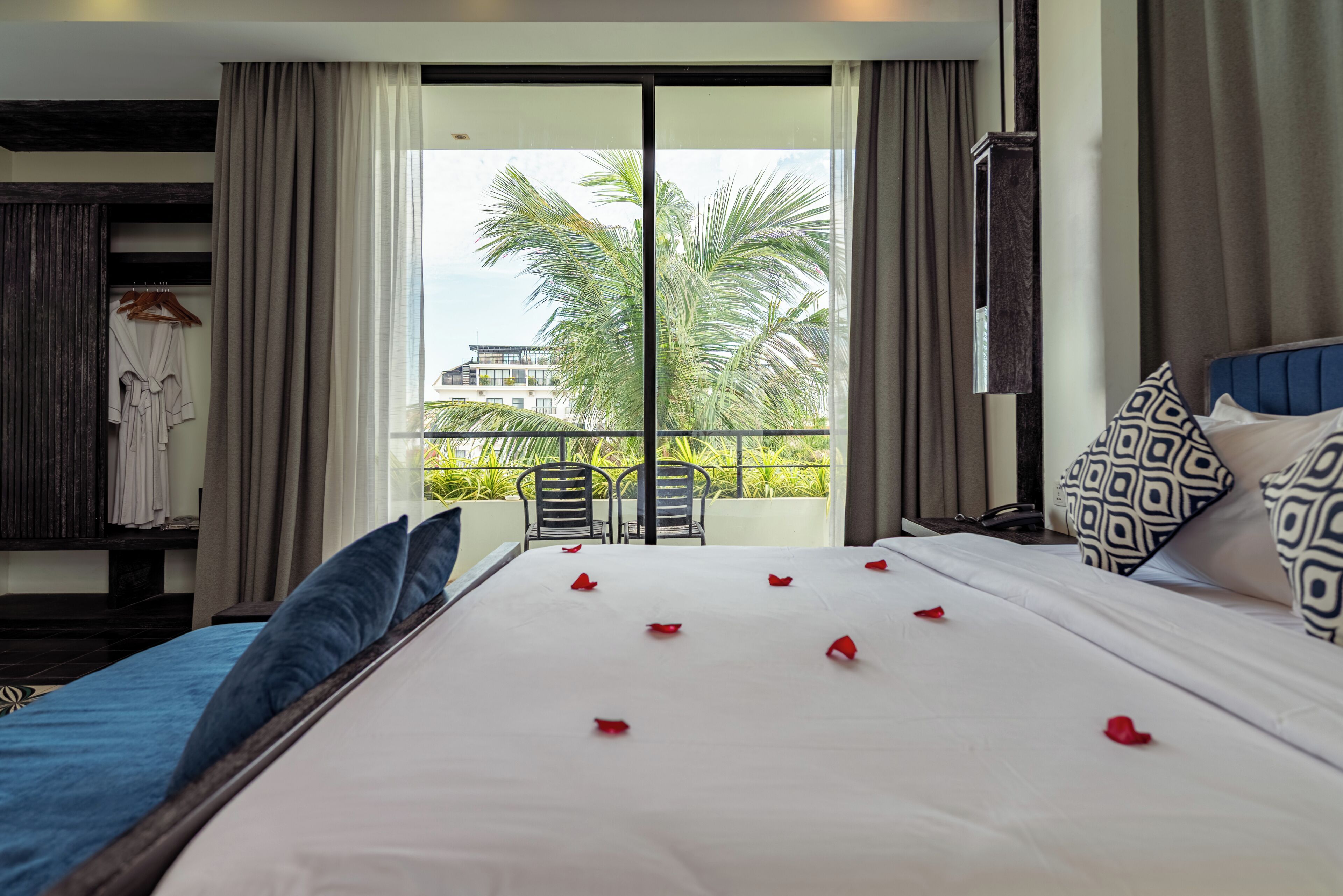 A hotel room with a large bed, a balcony with chairs, and a view of palm trees.